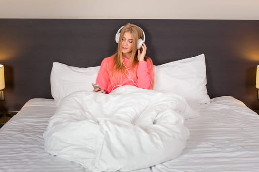Young pretty blond women smiling listening to the music with a headset from a smartphone lying on the bed at home