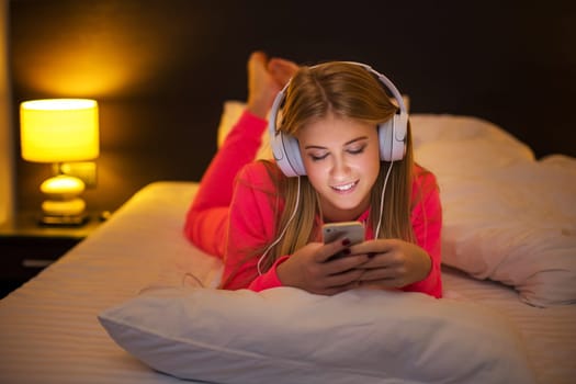 Young pretty blond women smiling listening to the music with headset from a smartphone lying on the bed at home