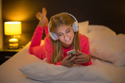 Young pretty blond women smiling listening to the music with a headset from a smartphone lying on the bed at home