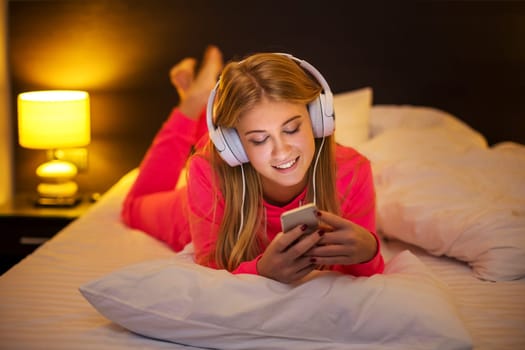 Young pretty blond women smiling listening to the music with headset from a smartphone lying on the bed at home