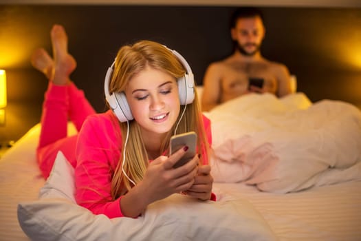 Young happy women on white bed using mobile phone with headphones, boyfriend on background. Concept about technology and people