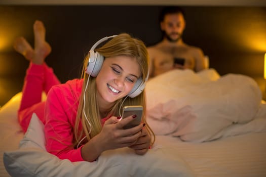 Young happy women on white bed using mobile phone with headphones, boyfriend on background. Concept about technology and people