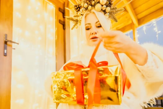 A woman in a white dress is holding a gold box with a red ribbon. She is wearing a crown on her head. The scene takes place in a room with a door and a window. The woman appears to be opening the gift box.