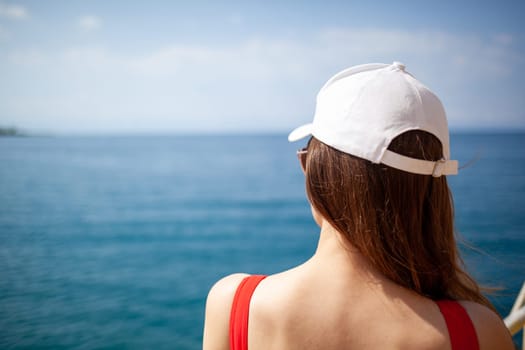 A beautiful girl in a red swimsuit with long hair, glasses and a white cap looks at the blue sea.