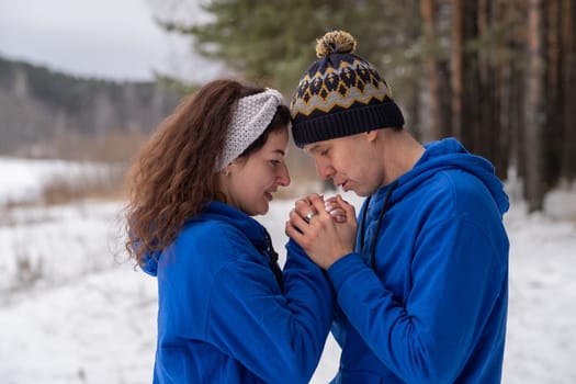 Outdoor happy couple in love posing in cold winter weather. A man and a woman in blue hoodies. Emotional young couple having fun while walking by winter forest, loving man hugging his laughing woman