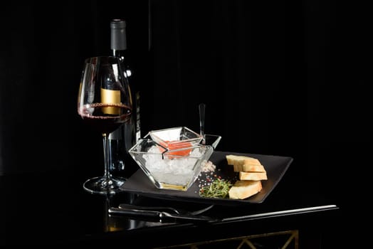 salmon fish caviar, on ice, with croutons and butter, on a transparent dish with a glass of red wine on a dark background