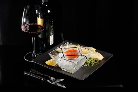 salmon fish caviar, on ice, with croutons and butter, on a transparent dish with a glass of red wine on a dark background
