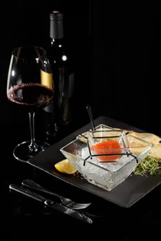 salmon fish caviar, on ice, with croutons and butter, on a transparent dish with a glass of red wine on a dark background