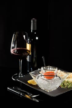 salmon fish caviar, on ice, with croutons and butter, on a transparent dish with a glass of red wine on a dark background