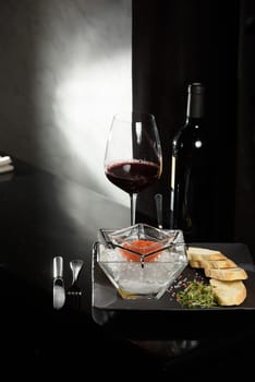 salmon fish caviar, on ice, with croutons and butter, on a transparent dish with a glass of red wine on a dark background