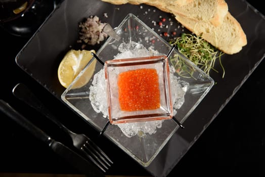 salmon fish caviar, on ice, with croutons and butter, on a transparent dish with a glass of red wine on a dark background