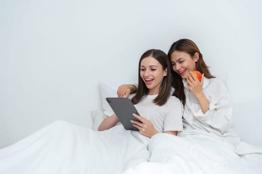 Young Asian lesbian couple sitting on bed during happy resting time, using tablet, exploring social media, chatting and relaxing in bedroom..