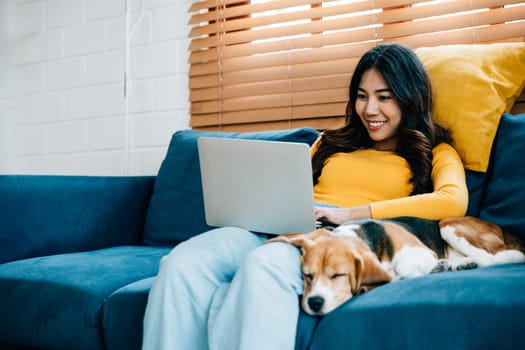 Working from her living room, a happy young woman shares the sofa with her laptop and Beagle dog. Her pet's peaceful sleep enhances the atmosphere of this friendly home office. Pet love