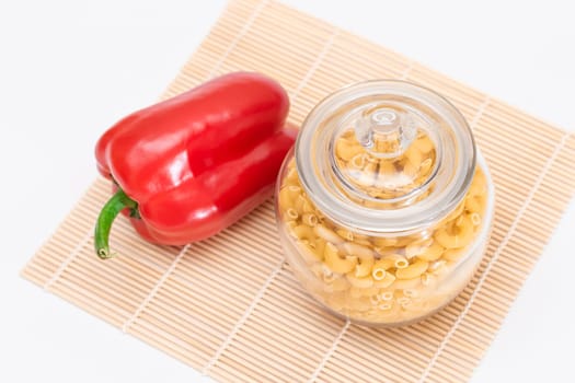 Uncooked Chifferi Rigati Pasta in Glass Jar and Red Bell Pepper on Bamboo Mat on White Background. Fat and Unhealthy Food. Classic Dry Macaroni. Italian Culture and Cuisine. Raw Pasta
