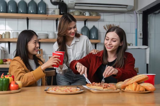 A group of young female friends have a party with pizza on the table and red drink glasses. Talk and live together happy, having fun at home..