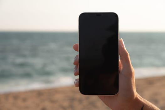 A girl holds a smartphone in her hand close-up with a black screen against the background of the sea. Technologies, modern methods of communication and remote work.
