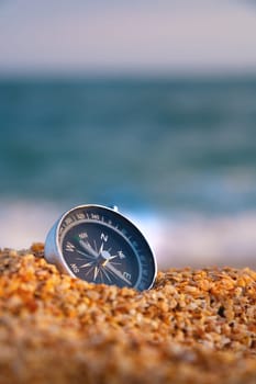 Conceptual photo, close-up of a compass lying in wet sand consisting of small crumbs of shells, against the backdrop of a blue calm sea.