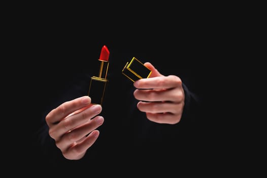 Hand holds red lipstick on black. Female hands opened an elite lipstick in a gold frame, serving a luxury cosmetic product.