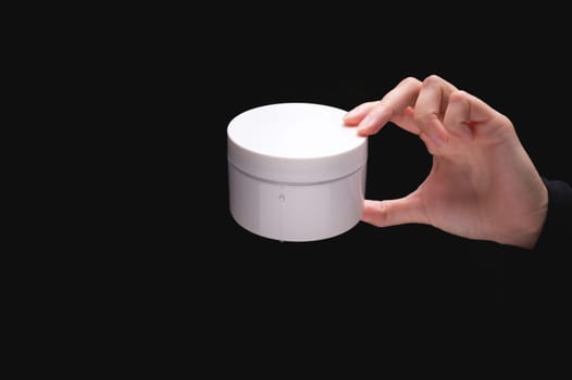 White cosmetic packaging mockup. A woman's hand holds a round jar of body cream on a black background. Beauty industry concept. Close-up.