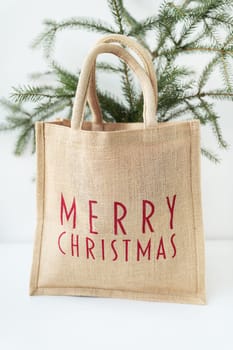 This is a photo of a burlap tote bag with Merry Christmas written in red on it, sitting in front of a small Christmas tree