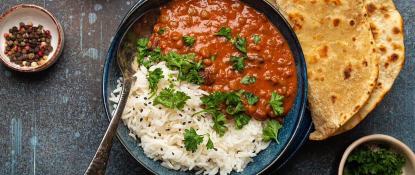 Traditional Indian Punjabi dish Dal makhani with lentils and beans in black bowl served with basmati rice, naan flat bread, fresh cilantro and spoon on blue concrete rustic table top view.