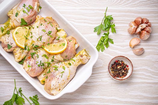 Raw uncooked chicken legs marinated with seasonings, herbs, lemon in white ceramic casserole top view on light wooden rustic background. Preparing healthy meal with marinated chicken drumsticks.