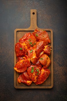 Raw uncooked chopped pieces of pork marinated with seasonings and parsley on wooden cutting board top view on dark rustic background. Cooking meal with marinated pork fillet pieces.