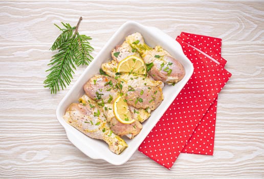 Preparing festive Christmas Eve meal with marinated chicken drumsticks. Raw chicken legs with herbs, lemon in white ceramic casserole top view on light wooden kitchen table with Christmas decoration.