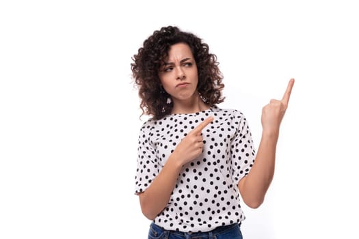 young authentic curly brunette slender lady wearing polka dot short sleeve shirt pointing fingers at ad space.
