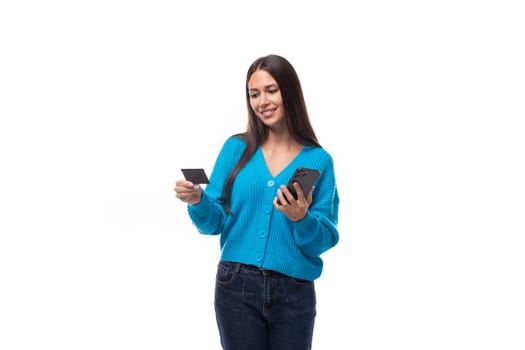 confused young brunette woman dressed in a blue sweater with buttons holds a bank card and a smartphone. e-business concept.