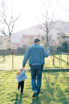 Dad leads a little girl by the hand through a green lawn to the house, looking at the phone. Back view. High quality photo