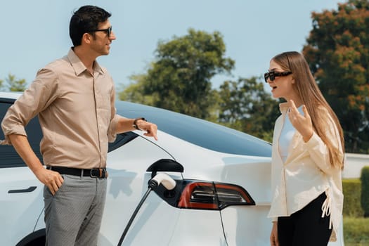 Young couple recharge electric car's battery from charging station in outdoor green city park in springtime. Rechargeable EV car for sustainable environmental friendly urban travel lifestyle.Expedient