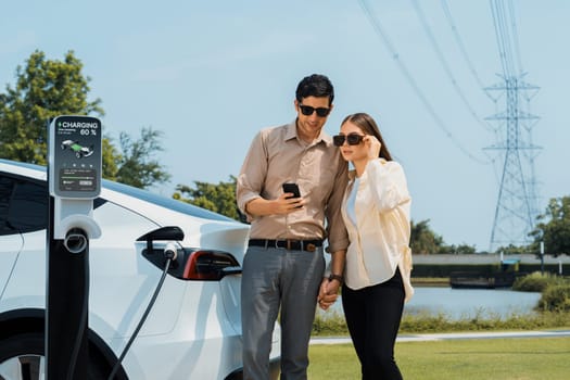 Couple pay for electricity with smartphone while recharge EV car battery at charging station connected to power grid tower electrical as electrical industry for eco friendly car utilization.Expedient