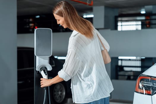 Young woman travel with EV electric car to shopping center parking lot charging in downtown city showing urban sustainability lifestyle by green clean rechargeable energy of electric vehicle innards