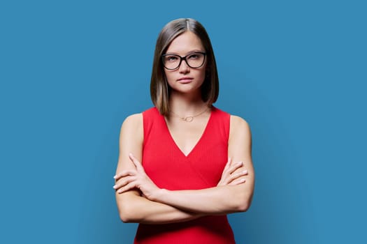 Young confident serious woman with eyeglasses in red on blue background. Successful fashionable female with crossed arms looking at camera. Business work services education, fashion beauty professions