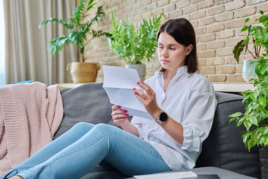 Serious young woman reading official paper letter while sitting on sofa in living room at home. Notification from bank, work, social tax services, invoice, insurance company, about rental property