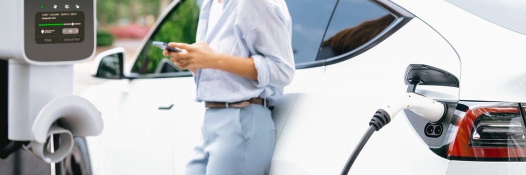 Young woman using smartphone online banking application to pay for electric car battery charging from EV charging station during vacation road trip at national park or summer forest. Panorama Exalt