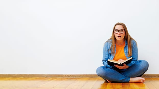 A surprised, positive young woman with glasses sits on the floor against a white wall holding a book