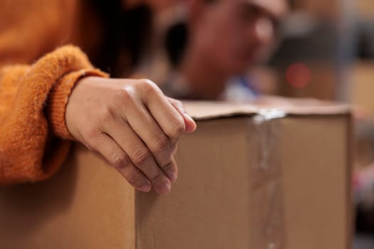 Package handler arm holding packed cardboard box in industrial warehouse. Storehouse employee picking order and preparing customer parcel for transportation with close up on worker hand