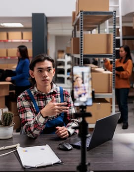 Asian man explaining parcel shipping for customer on smartphone camera in warehouse. Delivery manager working on laptop at storage room desk and recording video on mobile phone