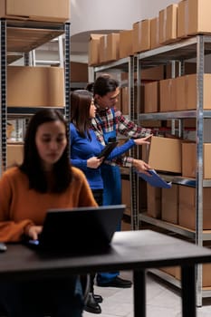 Postal storehouse employees managing order picking and shipping operation. Postal warehouse asian coworkers taking cardboard box from shelf and analyzing product checklist on digital tablet