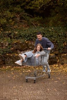 Autumn Joyride: Boy Rolling Girl in Shopping Cart amid Nature's Beauty. playful couple, autumnal adventure, outdoor fun, seasonal joy, laughter and love, romantic play, nature's embrace, joyful moments, couple goals, autumn magic.