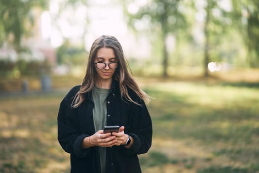 Young woman with glasses reading a message on her phone while standing in the park. High quality photo