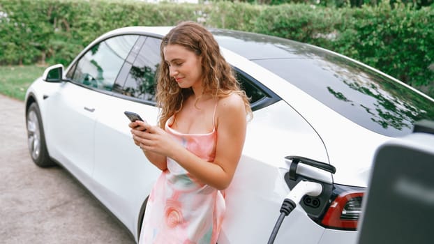 Young woman use smartphone to pay for electricity at public EV car charging station in nature. Modern environmental and sustainable automobile transportation lifestyle with EV vehicle. Synchronos