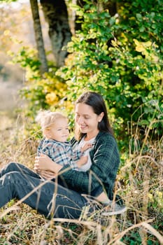 Smiling little girl sitting on lap of mother embracing her in park. High quality photo