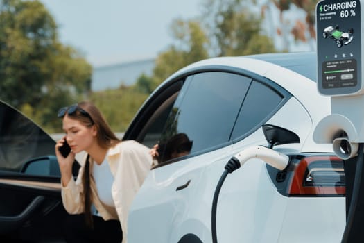 Young woman recharge her EV electric vehicle at green city park parking lot while talking on phone. Sustainable urban lifestyle for environment friendly EV car with battery charging station. Expedient