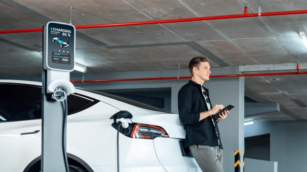 Young man travel with EV electric car to shopping center parking lot charging in downtown city showing urban sustainability lifestyle by green clean rechargeable energy of electric vehicle innards