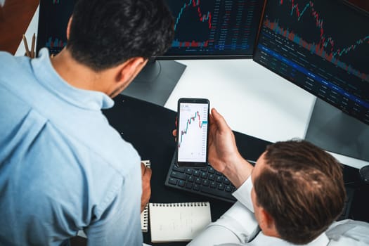 Investor stock officer focusing on dynamic exchange rate database on smartphone, comparing with market dynamic graph with monitor screen. Concept of analysis trading technology investment. Sellable.