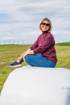 A middle-aged woman exuding charm and confidence, reclining gracefully on a haystack in a serene rural field, exuding an air of tranquility and natural beauty.