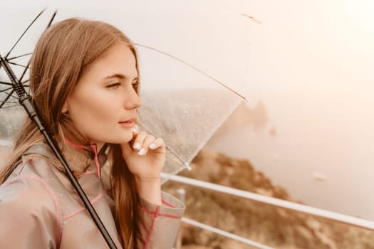 Woman rain park. Happy woman portrait wearing a raincoat with transparent umbrella outdoors on rainy day in park near sea. Girl on the nature on rainy overcast day